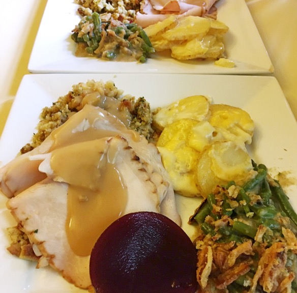 Two square white plates upon which are served thin slices of turkey, potatoes au gratin, green bean casserole, and bread stuffing covered in gravy. A standard American Thanksgiving meal.