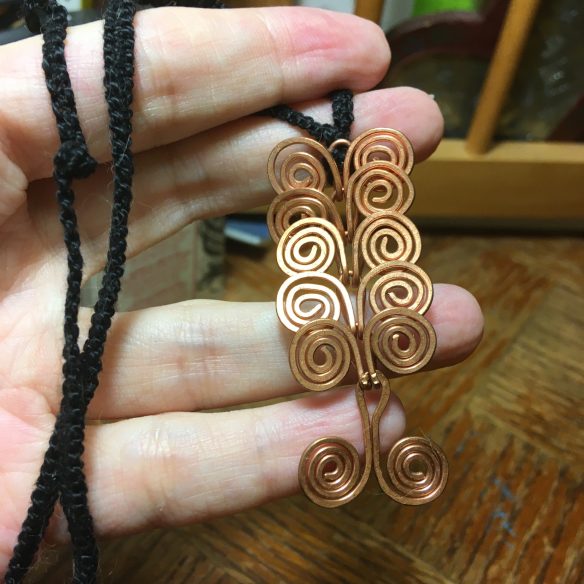 A woman's hand holds a pendant comprised of six  coiled copper wire links, each link having a flattened coil on each side. The pendant hangs from a black braided cord.
