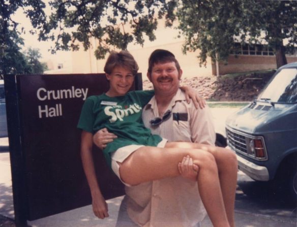 A heavy set man with short ginger hair and a bushy mustache carrying a petite slender girl with short blonde hair. Both are smiling broadly. They stand in front of a dorm building sign saying Crumley Hall. 