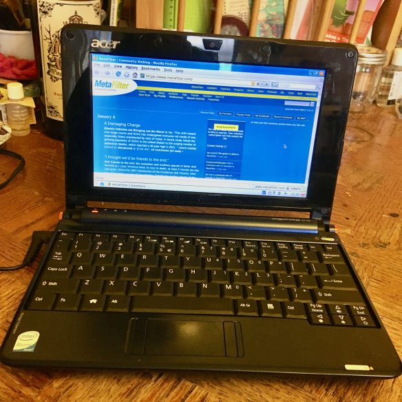 An open Acer Aspire One netbook, dark blue and sitting on a cluttered wooden table. On its screen can be seen the Metafilter webpage.