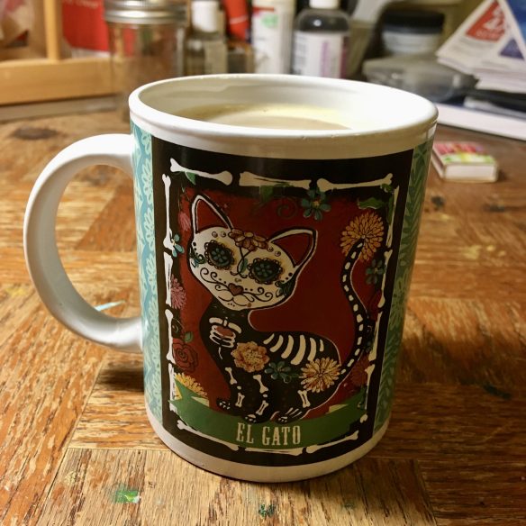 A mug filled to the brim with coffee sits on a cluttered wooden table surrounded by art supplies. On the mug an illustration of a skeleton cat in the style Dia de Meurtos (Day of the Dead) art. 