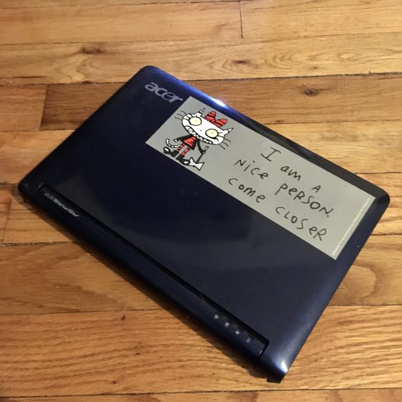 A closed Acer Aspire One netbook, dark blue, lying on a wooden floor. A sticker on it reads I am a nice person, come closer, and depicts a deranged cartoon cat holding an axe.