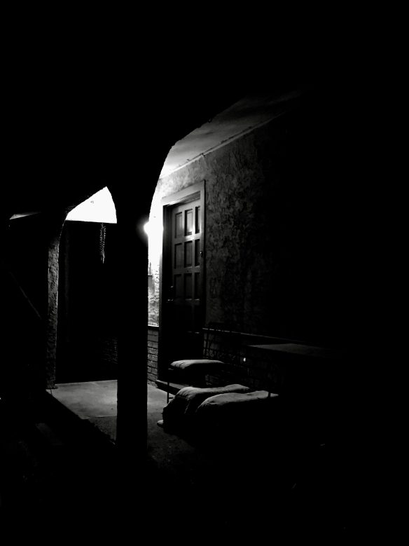A front porch of a small working class house at night. A single lightbulb dimly lights the area. 