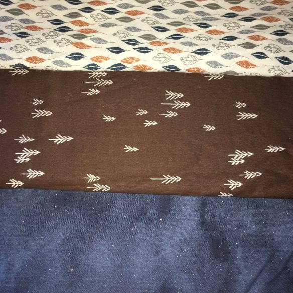 Three cuts of fabric stacked atop each other.  The top piece has a light beige background with alternating rows of dark teal, golden orange, and pale grey leaves. The middle piece has a milk chocolate background with a smattering of stick figure like pine trees sprinkled on it. The bottom piece is denim blue and has subtle little silver sparkles throughout.