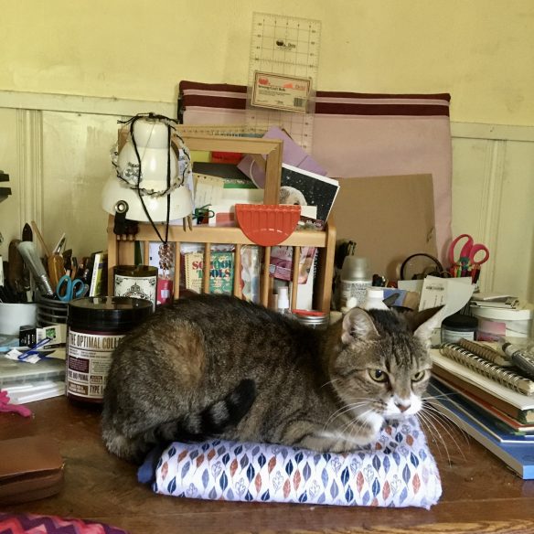 A small brown, black, and white tabby cat sits atop a stack of colorful fabrics on a messy table cluttered with all manner of arts and crafts supplies. 